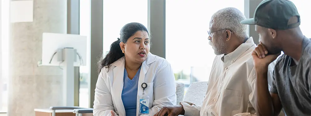 A female doctor in a white coat discusses the outpatient prospective payment system with an older man and a younger man in a waiting area, while large windows brighten the space. Both men appear to be listening attentively to her explanation.