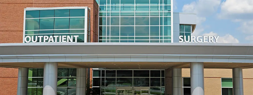 A modern medical building with large windows and a sign above the entrance that reads Outpatient Surgery in white letters, featuring PRO-PM technology. The sky is partly cloudy in the background.