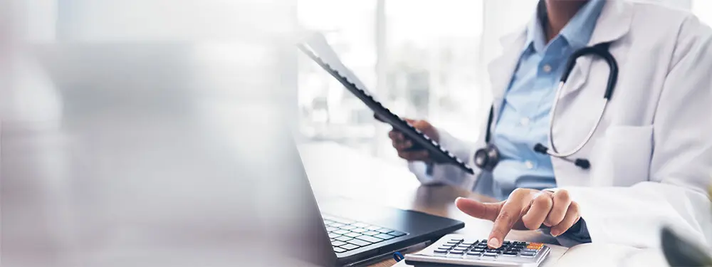 A person in a white lab coat with a stethoscope uses a calculator while holding a clipboard, sitting at a desk with a laptop, reviewing details of the CY 2025 OPPS and ASC Final Rule.