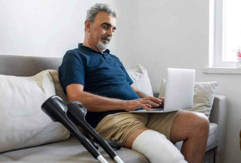 A man with a bandaged leg sits on a sofa using a laptop. Crutches rest beside him as he appears focused on work, possibly tracking patient-reported outcomes in orthopedics by the natural light coming through a nearby window.