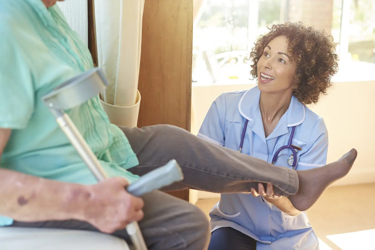 A nurse in scrubs kneels beside an elderly patient holding a crutch, gently supporting and examining the patient’s leg in a sunlit room, focusing on KCCQ heart failure reporting to improve care quality.