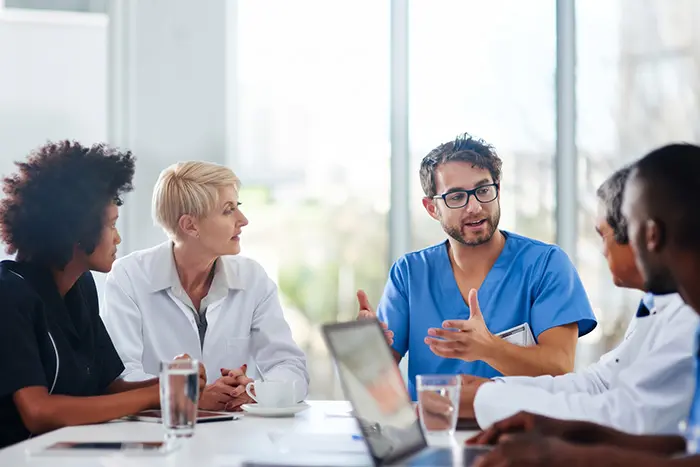 Four medical professionals in conversation at a table in a bright room, discussing physician peer review as one speaks in blue scrubs while others listen attentively. A laptop and glasses of water are on the table.