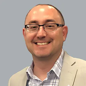 Matt Civili A smiling man with glasses, short brown hair, wearing a light gray blazer over a plaid shirt, poses in front of a plain gray background, representing CJR-X and strong physician partnerships.
